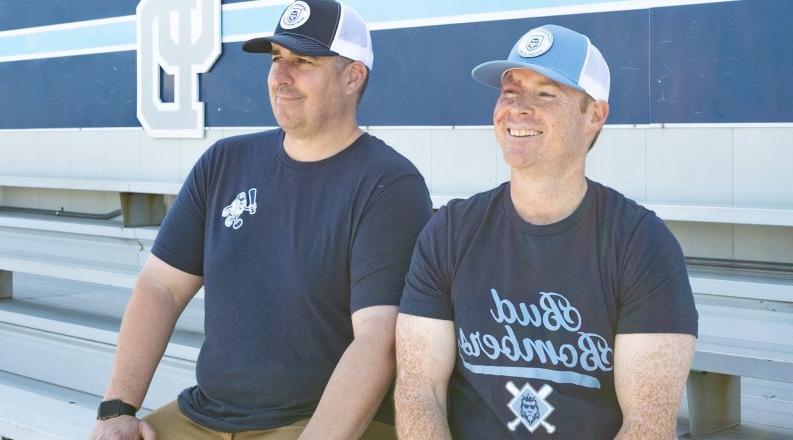 two men in blue shirts and white and blue caps sit on bleachers